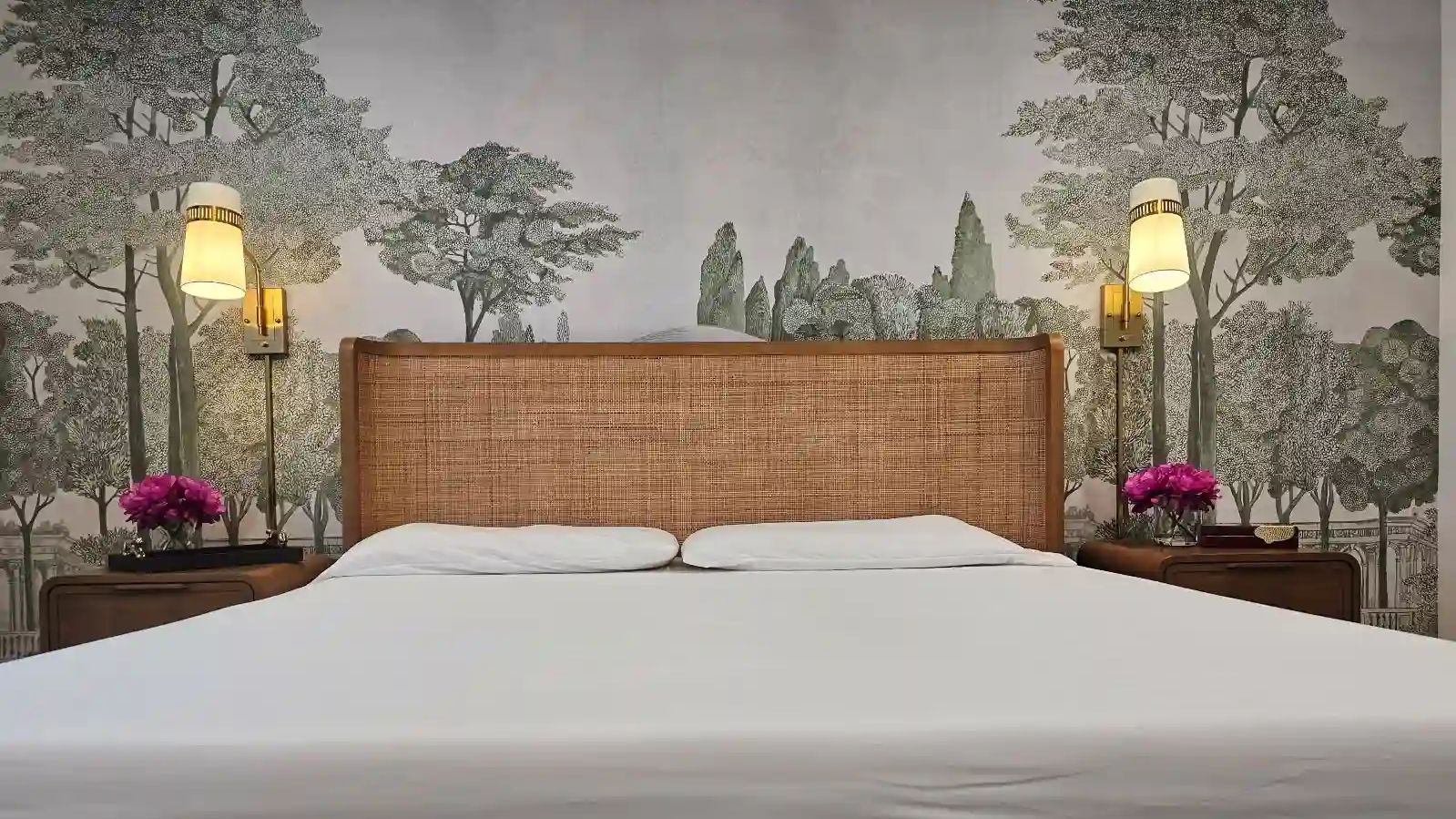 Bedroom with a misty forest wallpaper mural, a woven rattan headboard and two brass wall sconces